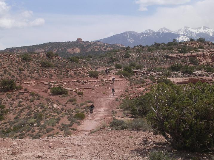 A scenic view of a rugged landscape featuring a dirt trail winding through a rocky area, with several hikers in the distance. The background showcases snow-capped mountains under a partly cloudy sky, surrounded by sparse vegetation and bushes typical of a desert environment. Porcupine Rim mountain bike trail.