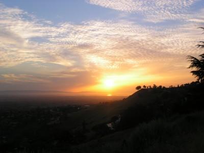 Sunset view over rolling hills, with vibrant orange and yellow hues reflecting off the horizon, and fluffy clouds scattered across a blue sky. Silhouetted trees dot the landscape in the foreground, creating a tranquil and picturesque scene. Alum Rock County Park mountain bike trail.