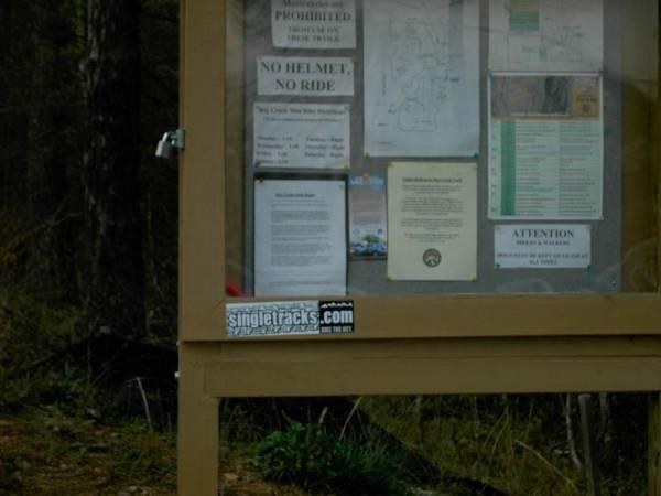 A wooden information board in a wooded area displaying various notices and maps for outdoor activities. The board includes a "No Helmet, No Ride" warning sign, informational flyers, and a sticker for a website related to singletrack trails. The surrounding environment features greenery and trees. Big Creek mountain bike trail.