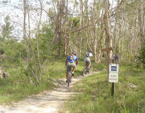 Three mountain bikers riding along a narrow dirt trail through a wooded area. A sign nearby indicates that helmets are mandatory for riders. The scene features sparse trees and greenery, with a clear path ahead. Markham Park mountain bike trail.