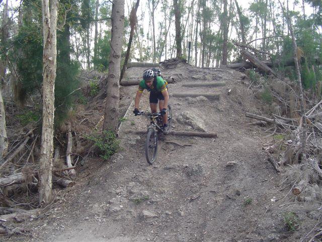 A mountain biker skillfully navigating a steep, rugged trail surrounded by trees and vegetation. The cyclist is in motion, descending a dirt slope with natural obstacles like roots and rocks. Markham Park mountain bike trail.