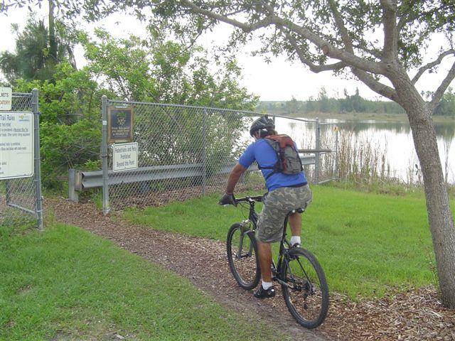 A person riding a mountain bike on a dirt path leading to a fenced area near a lake. Green vegetation and trees surround the pathway, and there is a signpost visible along the fence. Markham Park mountain bike trail.