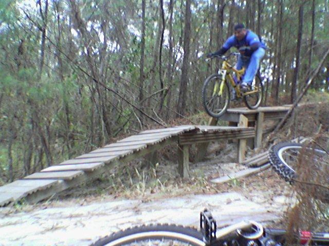 A person performing a jump on a yellow mountain bike over a wooden ramp in a wooded area, surrounded by trees and foliage. A second bike is partially visible in the foreground. Tillie Fowler Regional Park mountain bike trail.