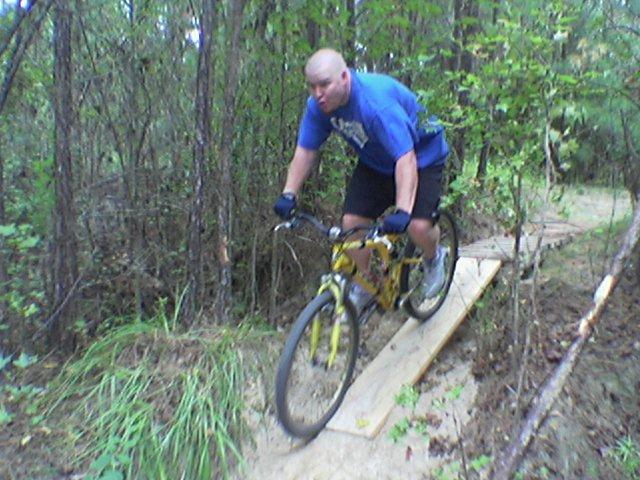 A person riding a yellow mountain bike on a wooden ramp through a wooded area, surrounded by trees and greenery. The rider is wearing a blue shirt and black shorts, focused on navigating the trail. Tillie Fowler Regional Park mountain bike trail.