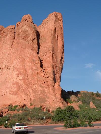 A tall, reddish rock formation rises against a clear blue sky, with green vegetation in the foreground and a parked white car at the base. Garden of the Gods: Ute Trail mountain bike trail.