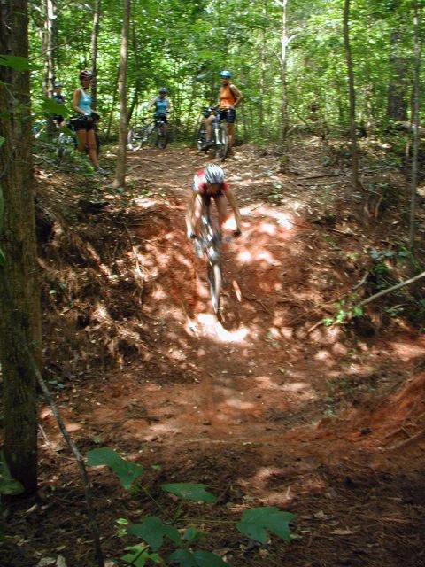 A mountain biker descends a dirt ramp in a wooded area with several trees. In the background, a few other cyclists are watching and waiting beside their bikes. The scene captures the thrill of mountain biking in nature. Fort Yargo State Park mountain bike trail.