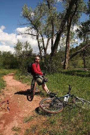 A person sitting on a bike path in a natural setting, wearing a red long-sleeve shirt and shorts. A bicycle lies on the ground nearby, surrounded by green grass and trees under a clear blue sky. Garden of the Gods: Ute Trail mountain bike trail.