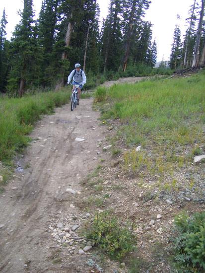 A mountain biker riding along a dirt path in a grassy area surrounded by tall evergreen trees, with a cloudy sky overhead. Keystone Resort Bike Park mountain bike trail.