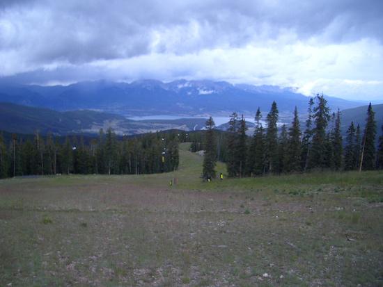 A panoramic view of a mountainous landscape, featuring green meadows, tall pine trees, and a distant lake surrounded by rolling hills. The sky is overcast with dark clouds, hinting at an impending storm. Keystone Resort Bike Park mountain bike trail.