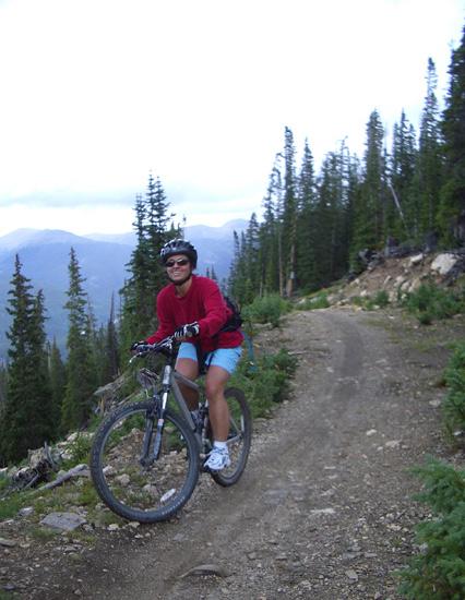 A person riding a mountain bike on a rugged dirt trail surrounded by tall pine trees and mountains in the background. The individual is wearing sunglasses, a red sweatshirt, and blue shorts, and is positioned beside the bike, smiling at the camera. The scene captures a sense of adventure and outdoor activity. Keystone Resort Bike Park mountain bike trail.