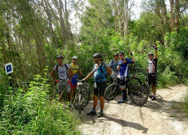 A group of six mountain bikers standing on a dirt path surrounded by lush greenery. They are wearing cycling gear and helmets, smiling for the camera, with their bikes beside them. Some members are raising their hands in a gesture of celebration. In the background, tall trees and bushes create a natural setting. Markham Park mountain bike trail.