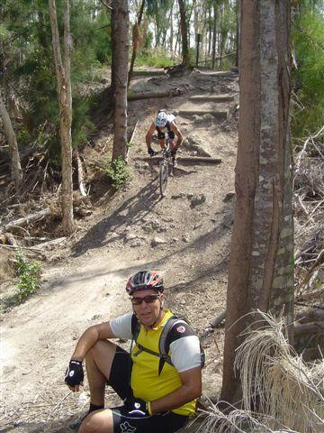 A mountain biker navigating a steep dirt trail lined with trees, while another biker rests on the ground in the foreground, wearing a helmet and cycling gear. Markham Park mountain bike trail.