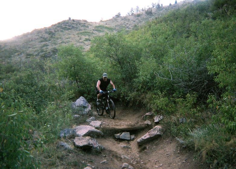 A mountain biker navigating a rocky trail through lush greenery, with hills visible in the background. The rider is wearing a sleeveless shirt and a cap, focused on maneuvering around large rocks on the path. Apex Park mountain bike trail.