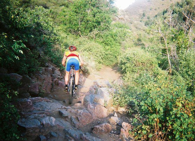 A person riding a mountain bike on a rocky, uneven trail surrounded by lush greenery and trees. The cyclist is wearing a bright red helmet and a colorful jersey, navigating through the outdoor terrain. Apex Park mountain bike trail.