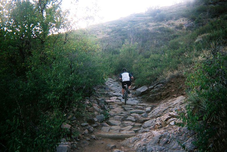 A mountain biker riding up a rocky trail surrounded by greenery, under a bright sky. The path is uneven and consists of large stones, leading toward a gently sloping hill in the distance. Apex Park mountain bike trail.