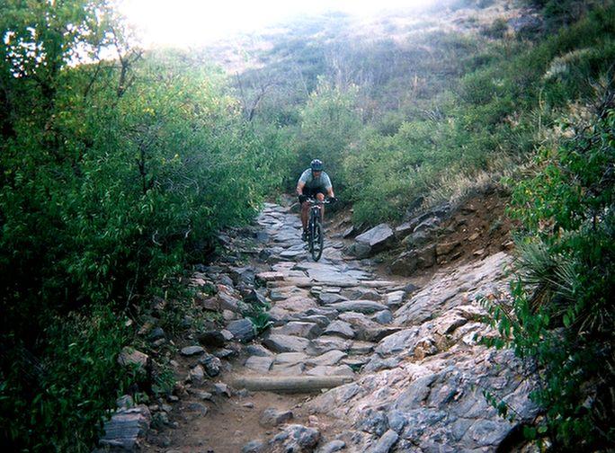 A mountain biker riding on a rocky trail surrounded by greenery and rocky terrain, going downhill on a sunny day. Apex Park mountain bike trail.