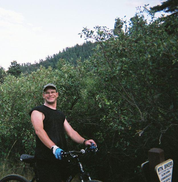 A smiling man in a sleeveless black shirt and blue gloves stands next to his mountain bike, surrounded by greenery and trees. A trail marker for mountain bikers is visible in the foreground. The sky is partly cloudy, indicating a pleasant outdoor setting. Apex Park mountain bike trail.