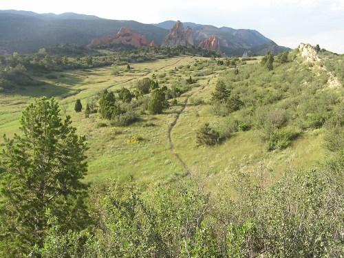 A scenic landscape featuring a wide green meadow with patches of trees and shrubs, leading towards rocky formations in the background under a clear blue sky. A winding path cuts through the grass, inviting exploration of the natural surroundings. Garden of the Gods: Ute Trail mountain bike trail.