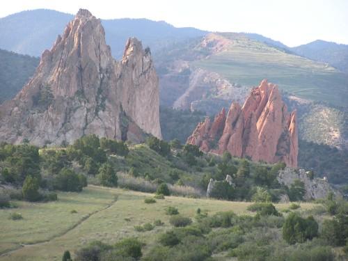 A scenic landscape featuring towering rock formations amidst lush greenery and rolling hills under a clear sky. The foreground displays rugged, reddish-brown rocks, while the background includes mountains and a grassy trail. Garden of the Gods: Ute Trail mountain bike trail.