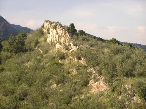 A rocky outcrop surrounded by lush greenery, with rolling hills and a distant mountain range under a partly cloudy sky. Garden of the Gods: Ute Trail mountain bike trail.