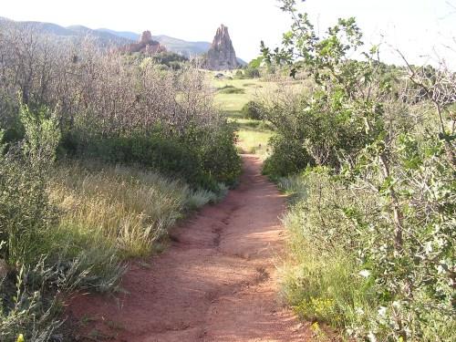 A dirt path winds through greenery, leading towards rocky formations and distant mountains under a clear sky. The area is surrounded by sparse bushes and tall grasses on either side. Garden of the Gods: Ute Trail mountain bike trail.