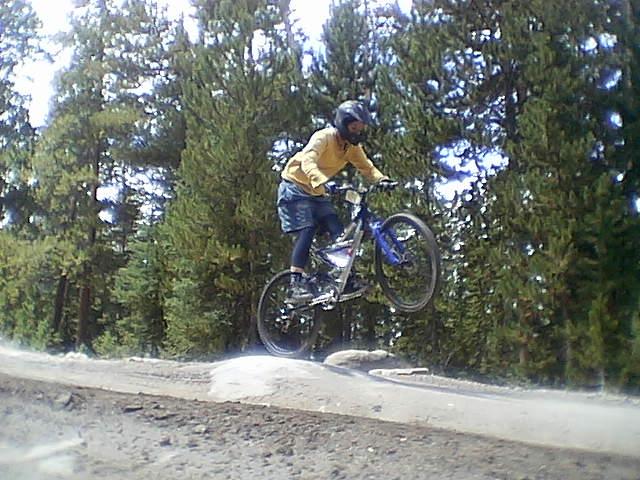 A person wearing a yellow long-sleeve shirt and a helmet is performing a bike jump on a dirt track surrounded by pine trees. The cyclist is airborne, with both wheels off the ground, showcasing an action shot of mountain biking. Keystone Resort Bike Park mountain bike trail.