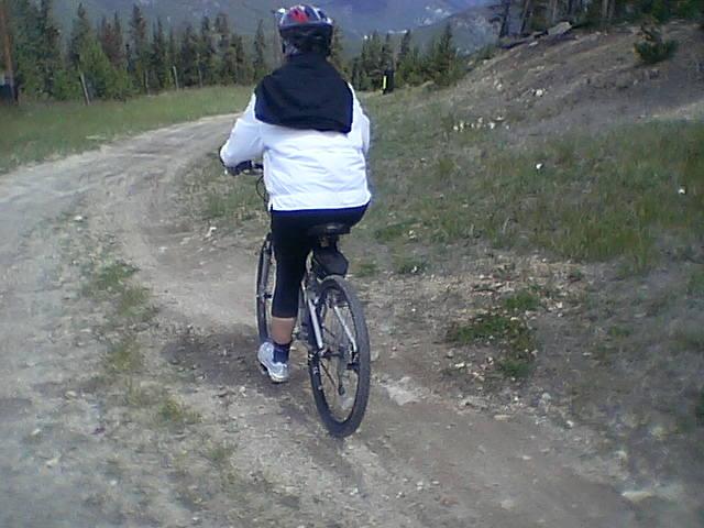 A person riding a mountain bike on a dirt trail surrounded by greenery and trees, wearing a white jacket and a helmet. The view is from behind, showing the cyclist navigating the path. Keystone Resort Bike Park mountain bike trail.