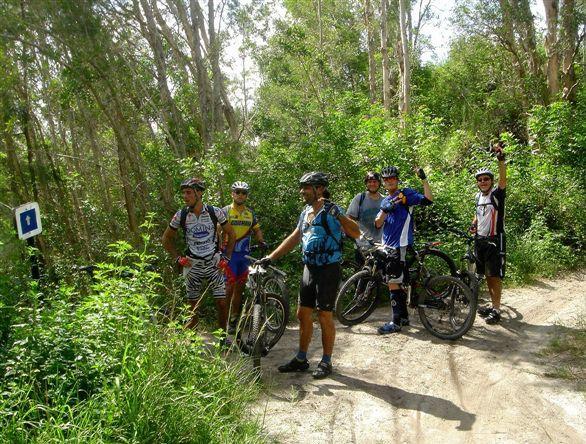 A group of six mountain bikers, wearing helmets and cycling gear, poses on a dirt trail surrounded by lush greenery and tall trees. Some members are holding their bikes while others are giving a thumbs up or waving at the camera, showcasing a sense of camaraderie and adventure. A trail sign is visible in the background. Markham Park mountain bike trail.