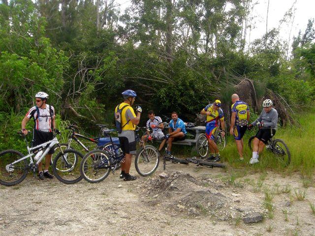 A group of cyclists taking a break in a wooded area, surrounded by trees and greenery. Some are standing near their bicycles, while others are seated at a picnic table. The scene captures a relaxed gathering of people in cycling gear, enjoying a moment outdoors. Markham Park mountain bike trail.