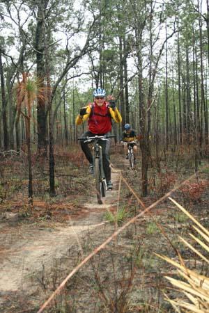 Two mountain bikers navigating a dirt trail through a forested area with tall trees. One biker is in the foreground, wearing a helmet and a red and yellow jacket, while the other is visible in the background. The landscape features a mix of greenery and dried vegetation, suggesting an outdoor adventure setting. Withlacoochee State Forest: Croom Section mountain bike trail.