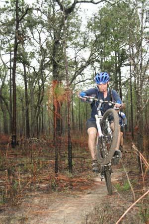 A mountain biker performing a wheelie on a dirt trail in a forested area, surrounded by trees and greenery. Withlacoochee State Forest: Croom Section mountain bike trail.