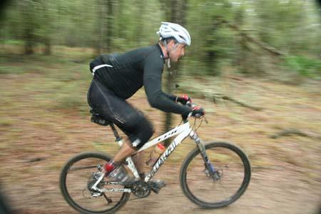 A cyclist in a black outfit and helmet rides a mountain bike through a wooded trail, showcasing motion with a blurred background to convey speed. Withlacoochee State Forest: Croom Section mountain bike trail.