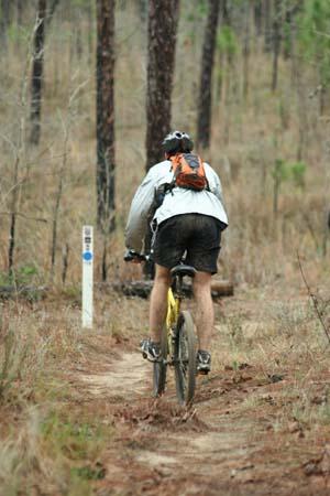 A person riding a mountain bike on a narrow trail surrounded by trees in a forested area. The cyclist is wearing a light jacket and shorts, and has a backpack on. A signpost is visible in the background. Withlacoochee State Forest: Croom Section mountain bike trail.