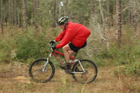 A person wearing a red jacket and a helmet is riding a mountain bike through a wooded area, with trees and underbrush visible in the background. Withlacoochee State Forest: Croom Section mountain bike trail.