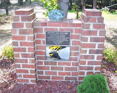 A brick monument with a plaque commemorating the site of "Fire Wise Park," surrounded by greenery and landscaping features. The plaque includes information about the park's purpose and history. Pee Wee's Mountain Bike Park mountain bike trail.