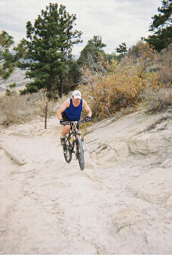 A mountain biker in a blue tank top and shorts navigates a rocky trail, surrounded by trees and shrubs under a cloudy sky. Red Rocks / Dakota Ridge mountain bike trail.