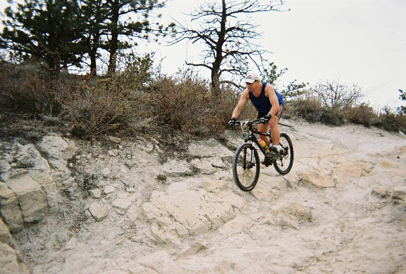 A cyclist descending a rocky trail surrounded by sparse vegetation and trees. The rider is wearing a blue tank top and shorts, concentrating on navigating the rugged terrain. The scene captures the excitement and challenge of mountain biking in a natural setting. Red Rocks / Dakota Ridge mountain bike trail.