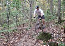 A mountain biker wearing a helmet jumps off a rocky ledge on a dirt trail, surrounded by trees with autumn foliage. Blankets Creek mountain bike trail.