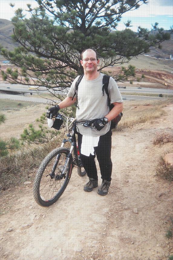 A person standing next to a mountain bike on a dirt trail surrounded by grassy terrain and a pine tree, wearing gloves and casual outdoor clothing. Red Rocks / Dakota Ridge mountain bike trail.