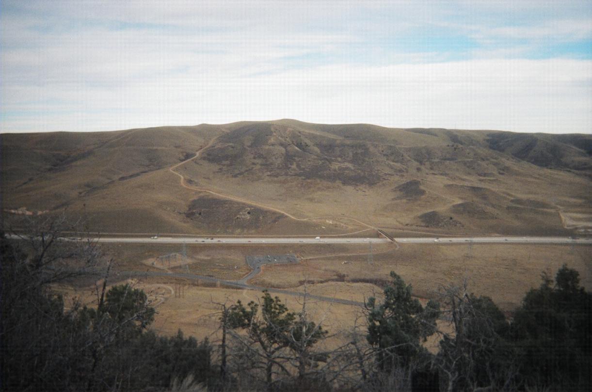 A scenic view of rolling hills under a cloudy sky, with a highway cutting through the landscape. Sparse vegetation is visible in the foreground, while dirt paths wind up the hills in the background. Red Rocks / Dakota Ridge mountain bike trail.