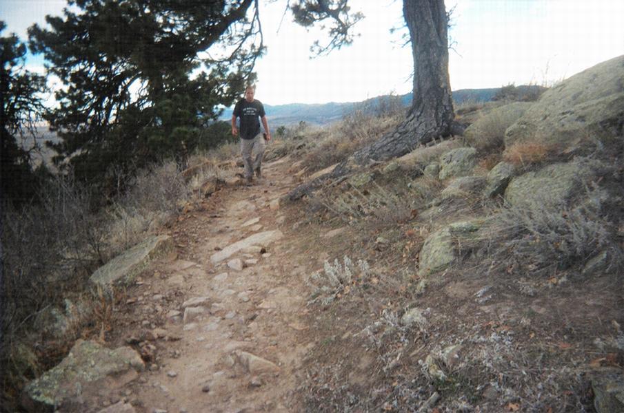 A person walking along a rocky hiking trail surrounded by trees and shrubs in a mountainous landscape. The scene captures the natural beauty of the outdoors with a clear sky in the background. Red Rocks / Dakota Ridge mountain bike trail.