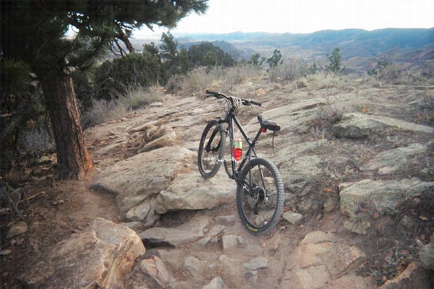 A mountain bike resting on rocky terrain with a scenic view of rolling hills and trees in the background. Red Rocks / Dakota Ridge mountain bike trail.