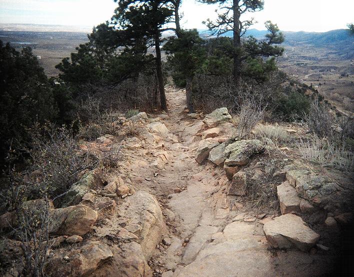 A rocky hiking trail surrounded by trees, winding along a hillside with a view of distant landscapes under a cloudy sky. Red Rocks / Dakota Ridge mountain bike trail.