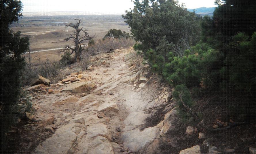 A rocky hiking trail winding through a sparse landscape, bordered by shrubs and trees, with a distant view of rolling hills and an expansive sky. Red Rocks / Dakota Ridge mountain bike trail.