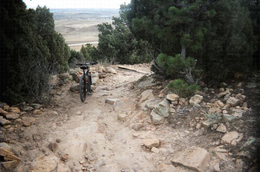 A mountain bike parked on a rocky trail surrounded by trees and shrubs, leading down a steep path with scenic views in the background. Red Rocks / Dakota Ridge mountain bike trail.
