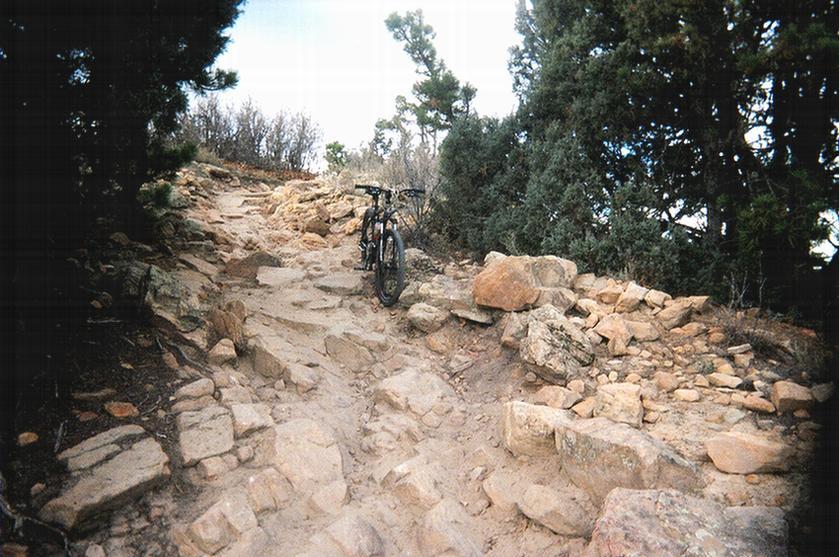 A mountain bike resting on a rocky trail surrounded by shrubs and trees, depicting a rugged outdoor landscape. Red Rocks / Dakota Ridge mountain bike trail.
