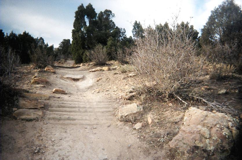A rugged dirt path winds through a landscape of low shrubs and scattered rocks, leading towards a tree line in a natural outdoor setting. The sky is overcast, adding a muted atmosphere to the scene. Red Rocks / Dakota Ridge mountain bike trail.