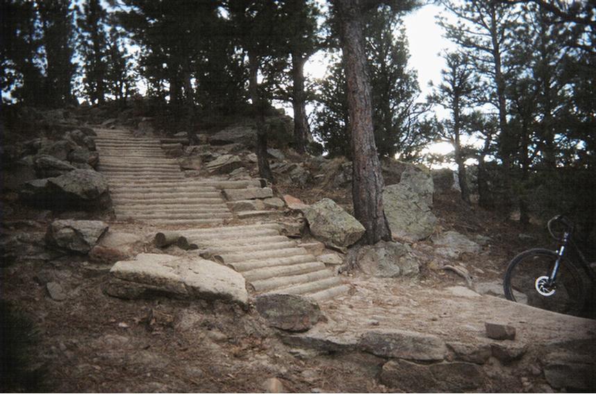 A rocky staircase leading uphill through a forested area, surrounded by trees and boulders, with a mountain bike partially visible on the right side of the image. Red Rocks / Dakota Ridge mountain bike trail.