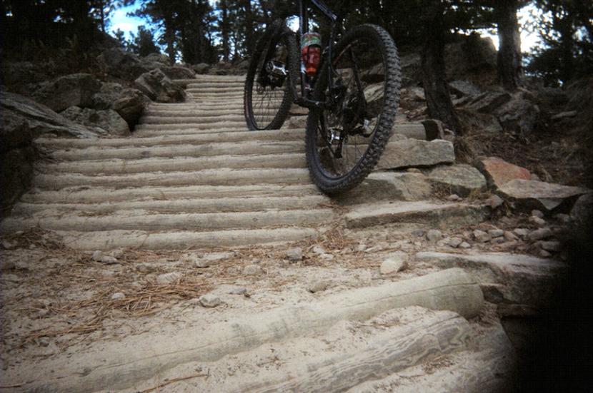 A close-up view of a mountain bike positioned near a set of wooden stairs on a trail, surrounded by rocky terrain and trees. The ground is sandy with scattered stones, and the sky is partially visible in the background. Red Rocks / Dakota Ridge mountain bike trail.
