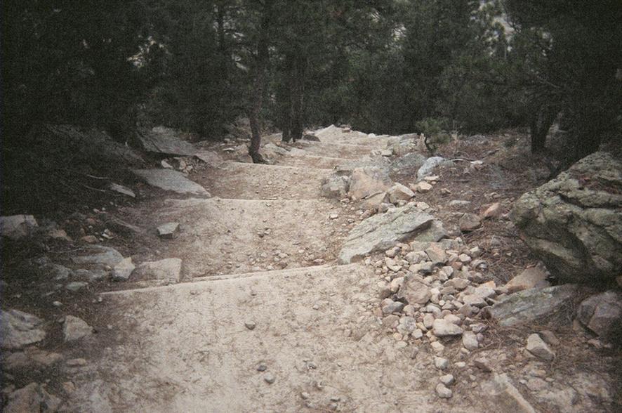 A narrow, rocky hiking trail winding through a forest, lined with trees on either side. The path is covered with dirt and stones, and gently ascends in the distance. Red Rocks / Dakota Ridge mountain bike trail.
